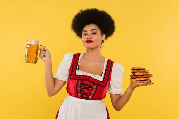 Felice afro americano oktoberfest cameriera in Dirndl tenendo boccale di birra e gustosi pretzel su giallo — Foto stock