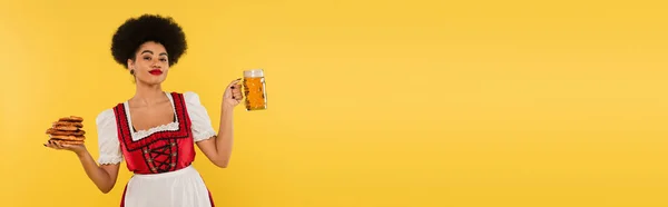 Serveuse bavaroise afro-américaine en dirndl tenant une tasse de bière et de savoureux bretzels sur jaune, bannière — Photo de stock