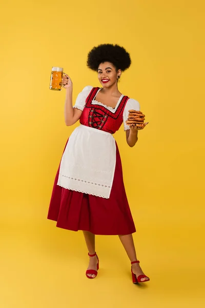 Cheerful african american bavarian waitress in dirndl holding beer mug and tasty pretzels on yellow — Stock Photo