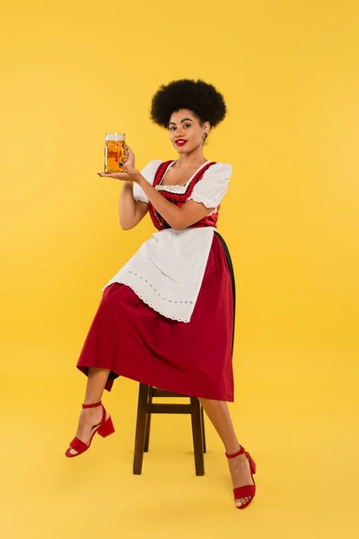 Jovem garçonete afro-americana em vestido bávaro autêntico sentado na cadeira com caneca de cerveja no amarelo — Fotografia de Stock