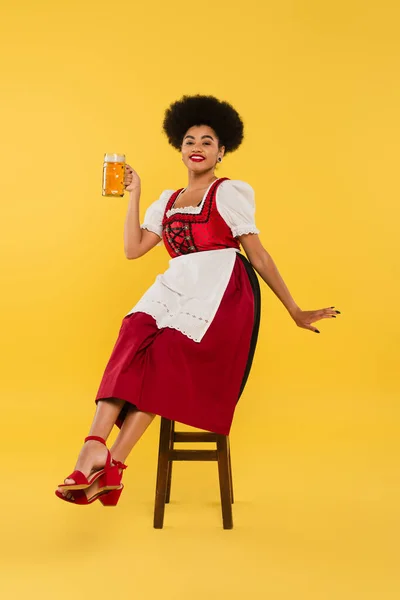 Joyous african american waitress in oktoberfest outfit sitting on chair with beer mug on yellow — Stock Photo