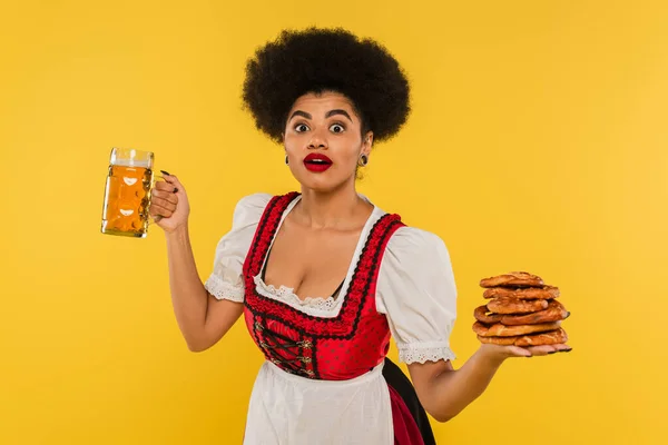 Amazed african american oktoberfest waitress with beer mug and pretzels looking at camera on yellow — Stock Photo