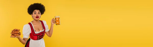 Удивленный африканский октоберфест официантка с кружкой пива и крендельками на желтом, баннер — стоковое фото