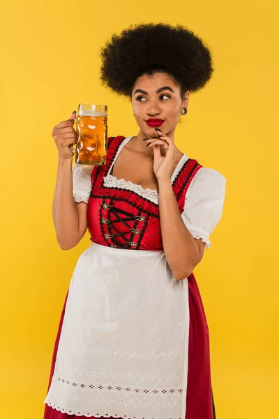 Difícil africano americano oktoberfest camarera en dirndl traje de pie con taza de cerveza en amarillo - foto de stock