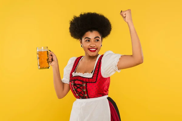 Ravie serveuse bavaroise afro-américaine avec tasse de bière montrant geste de victoire sur jaune — Photo de stock