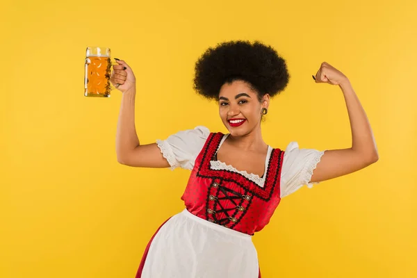 Cheerful african american oktoberfest waitress with beer mug showing triumph gesture on yellow — Stock Photo