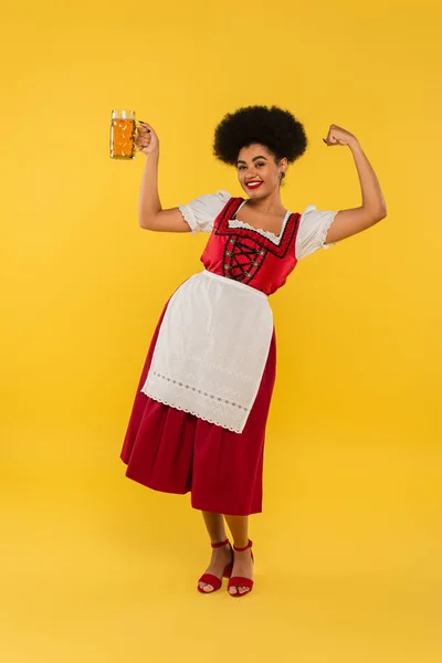 Aufgeregte afrikanisch-amerikanische Oktoberfest-Kellnerin mit Bierkrug zeigt Siegergeste auf gelb — Stockfoto