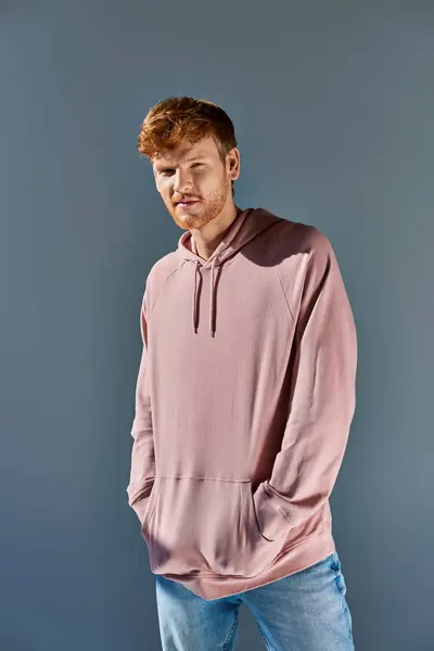Stylish young red haired man in pink hoodie and jeans looking at camera posing on grey backdrop — Stock Photo