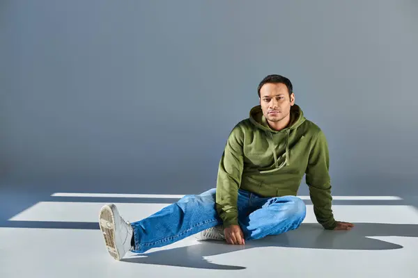 Schöner junger indischer Mann in khakifarbenem Kapuzenpulli und Jeans sitzt auf dem Boden und schaut in die Kamera — Stockfoto