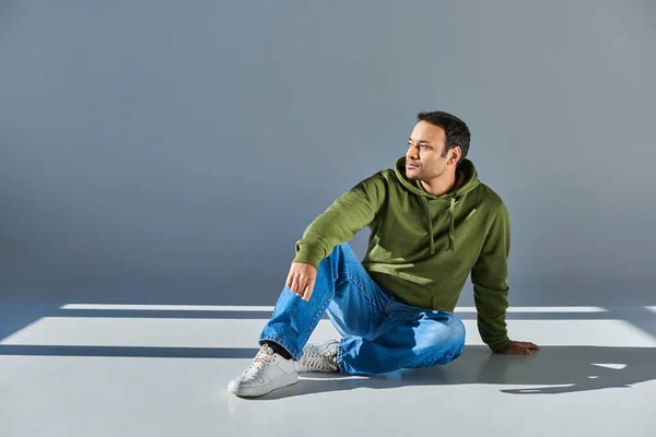 Indischer Mann in lässiger Streetwear sitzt auf dem Boden und schaut vor grauem Hintergrund weg — Stockfoto
