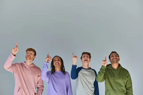 Four joyful men in street wear looking up and pointing up on grey background, cultural diversity — Stock Photo