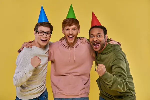 Trois amis gais multiculturels dans des chapeaux d'anniversaire étreignant et regardant la caméra, anniversaire — Photo de stock