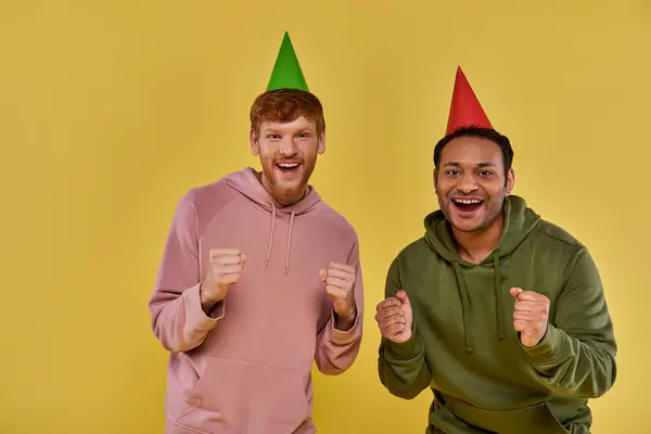 Close up two multiracial friends in casual outfits and birthday hats with fists up raised, birthday — Stock Photo