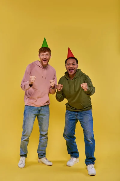 Dois homens multirraciais bonitos em chapéus de aniversário de pé e celebrando com punhos para cima, aniversário — Fotografia de Stock