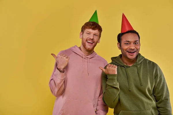 Dos jóvenes amigos en atuendo casual y sombreros de cumpleaños sonriendo a la cámara señalando los pulgares a un lado - foto de stock