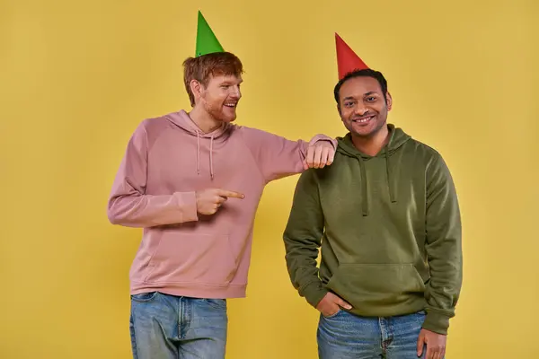 Dois homens alegres em trajes casuais e chapéus de aniversário posando juntos no fundo amarelo, aniversário — Fotografia de Stock