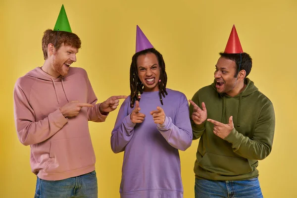 Três amigos multiculturais em chapéus de aniversário celebrando e apontando os dedos para a câmera, aniversário — Fotografia de Stock