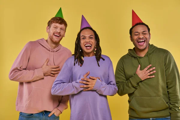 Young multiethnic friends in casual outfits and birthday hats laughing sincerely, yellow backdrop — Stock Photo