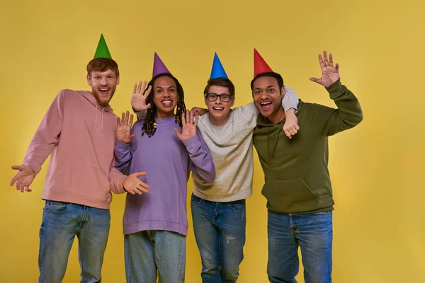 Four young friends in birthday hats smiling at camera waving and showing open palms, birthday — Stock Photo