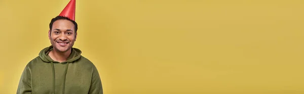 Улыбающийся индиец в толстовке цвета хаки и шляпе на день рождения, смотрящий в камеру на желтом фоне, день рождения — стоковое фото