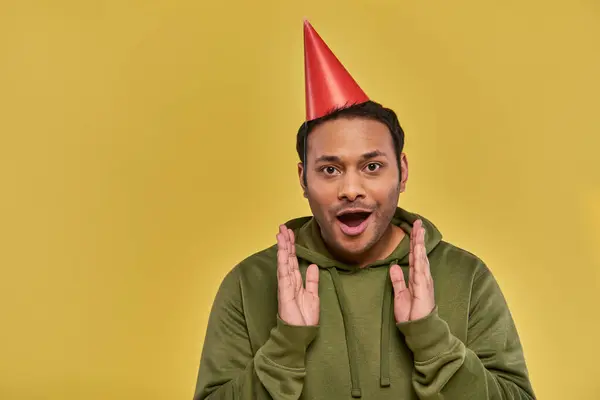 Astonished young indian man in birthday hat and khaki hoodie with hands closer to face, birthday — Stock Photo