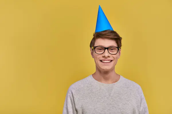 Jovem homem alegre em camisola branca e chapéu de aniversário sorrindo para a câmera no fundo amarelo — Fotografia de Stock