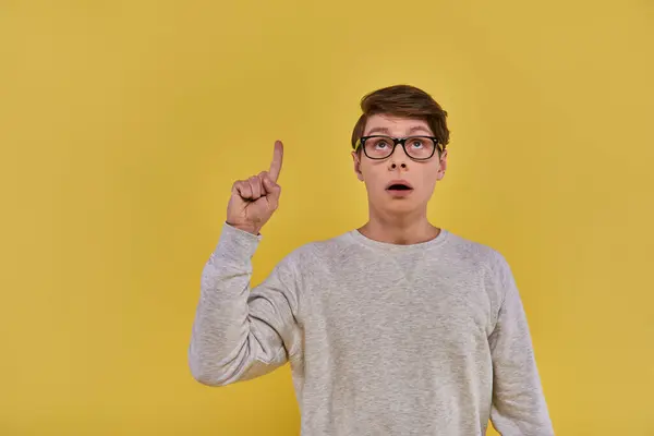 Young man in white sweatshirt raising his finger in eureka gesture and looking up on yellow backdrop — Stock Photo
