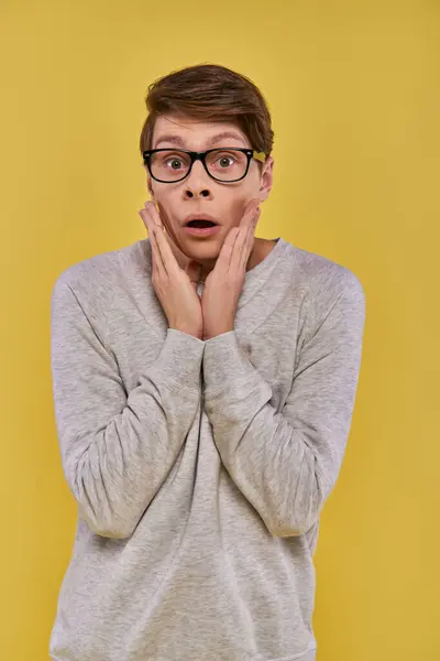 Shocked young man in casual attire with glasses touching his cheeks with hands on yellow backdrop — Stock Photo