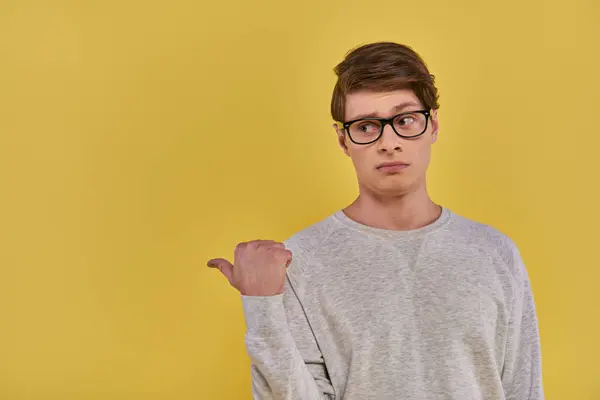 Young upset man in casual wear and glasses pointing finger aside on left side on yellow backdrop — Stock Photo
