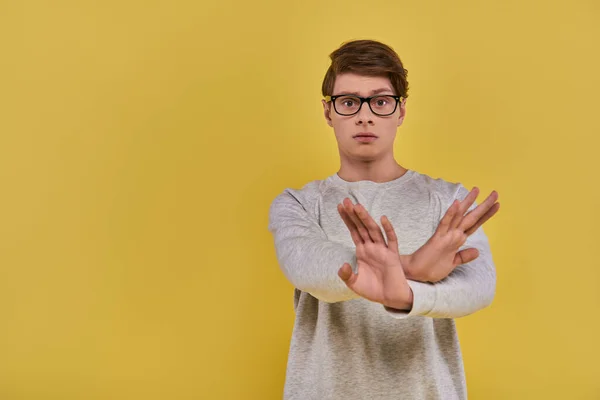 Jeune homme triste en sweat-shirt blanc avec des lunettes montrant le déni geste de la main croisée sur fond jaune — Photo de stock