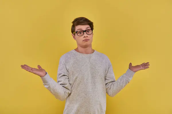 Jovem sem noção confuso em camisola e óculos mostrando gesto impotente no fundo amarelo — Fotografia de Stock