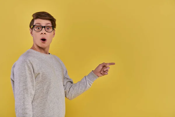 Erstaunt fröhlicher Mann in Freizeitkleidung mit Brille, Zeigefinger auf der rechten Seite, gelber Hintergrund — Stockfoto