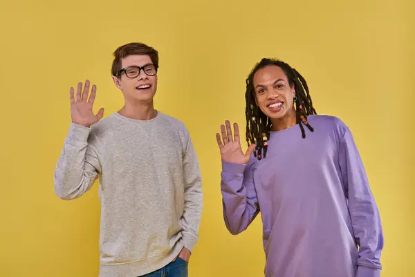 Two cheerful friends in purple and white sweatshirts waving and smiling at camera, diversity — Stock Photo