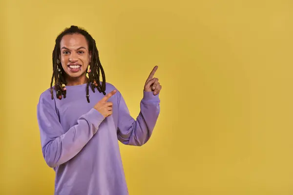 Cheerful african american man with dreadlocks pierced lip pointing on right side on yellow backdrop — Stock Photo