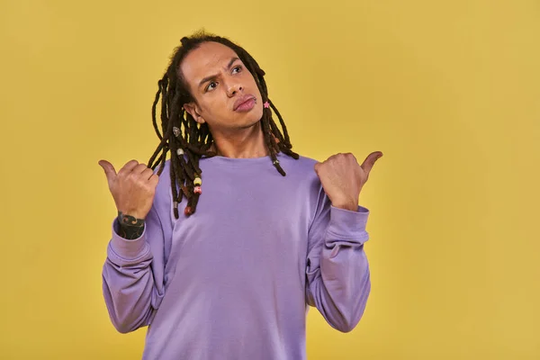 Confused and puzzled young man with his thumbs pointing different ways on yellow background — Stock Photo