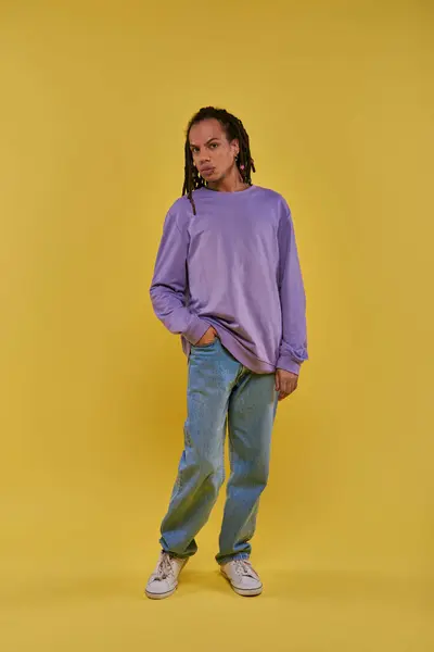 Joven afroamericano en sudadera púrpura y jeans con rastas posando sobre fondo amarillo - foto de stock