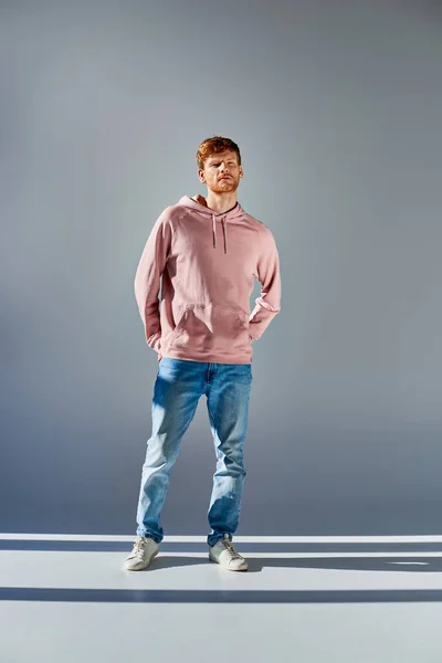 Bellissimo uomo dai capelli rossi in felpa rosa con cappuccio in posa con le mani in tasche di jeans su sfondo grigio — Foto stock