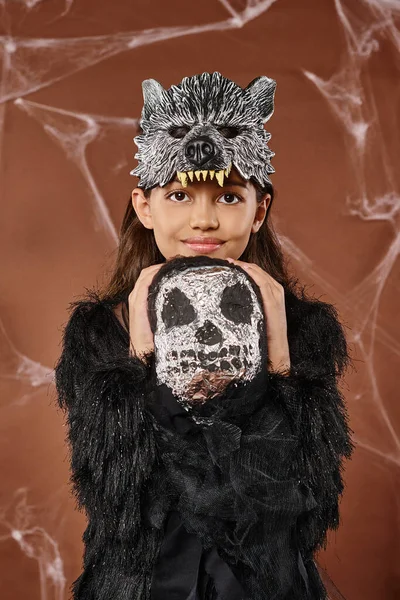 Portrait of smiling girl in wolf mask and black attire hugging spooky toy, Halloween, close up — Stock Photo