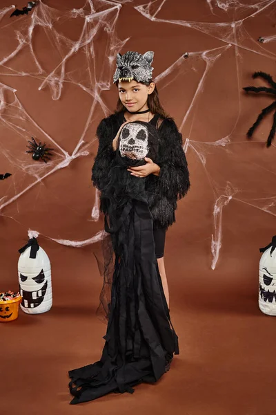 Cute preteen girl holds Halloween toy on brown backdrop with cobweb and spiders, Halloween concept — Stock Photo