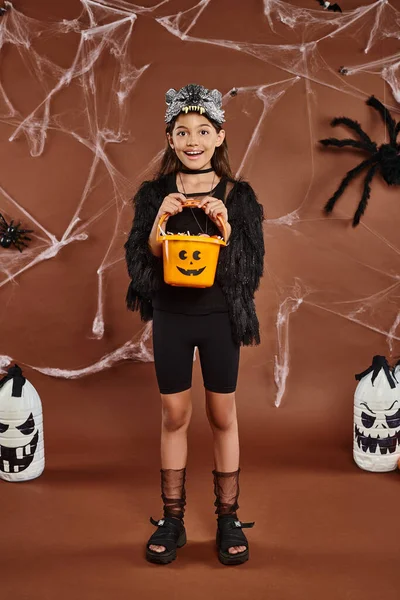 Chica feliz sosteniendo cubo de dulces en máscara de lobo y traje negro, concepto de Halloween - foto de stock