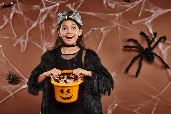 Chiudere allegra ragazza con il suo secchio pieno di dolci su sfondo marrone, concetto di Halloween — Foto stock