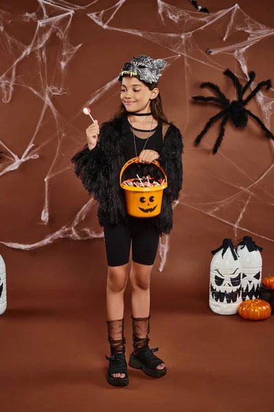 Glückliches preteen Mädchen, das Lutscher betrachtet und Eimer mit Süßigkeiten in der Hand hält, Halloween-Konzept — Stockfoto