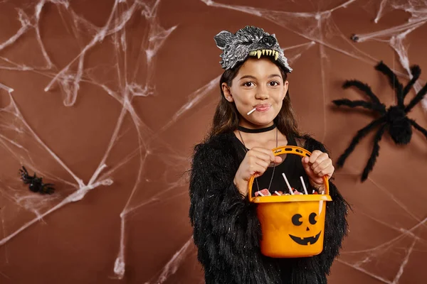Close up girl with lollipop holding candy bucket on brown backdrop with spiderweb, Halloween — Stock Photo