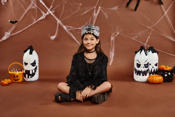 Cheerful preteen girl sitting with legs crossed on brown backdrop with lanterns and web, Halloween — Stock Photo