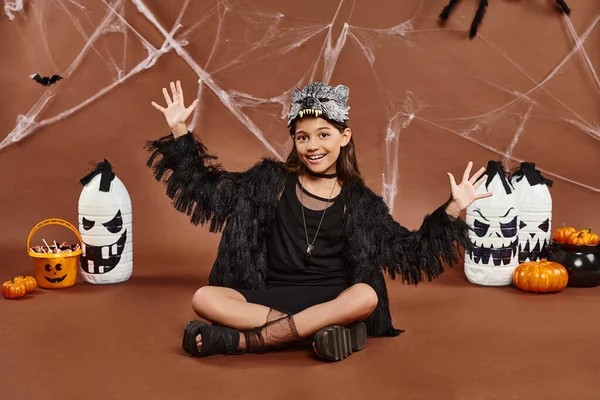 Happy girl in wolf mask and black attire sitting with legs crossed and showing open palms, Halloween — Stock Photo