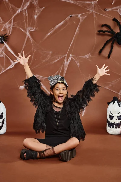 Close up cheerful girl yaying sitting with crossed legs on brown backdrop with spiderweb, Halloween — Stock Photo