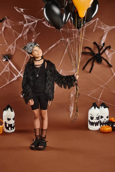 Sonriente chica sostiene negro y naranja globos en marrón telón de fondo con web y arañas, Halloween - foto de stock
