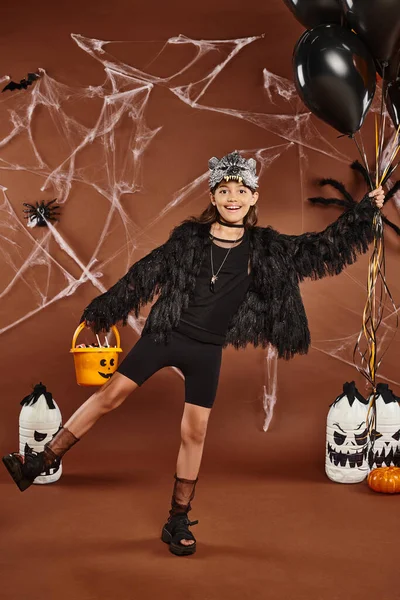 Chica feliz de pie en una pierna sostiene globos negros y naranjas y cubo con dulces, Halloween - foto de stock
