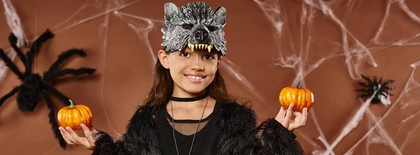 Preteen girl in wolf mask holds pumpkins on brown backdrop with spiders and web, Halloween, banner — Stock Photo