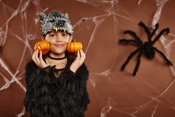 Cerca de niña sonriente en máscara de lobo sostiene calabazas cerca de su cara con araña en el telón de fondo, Halloween - foto de stock